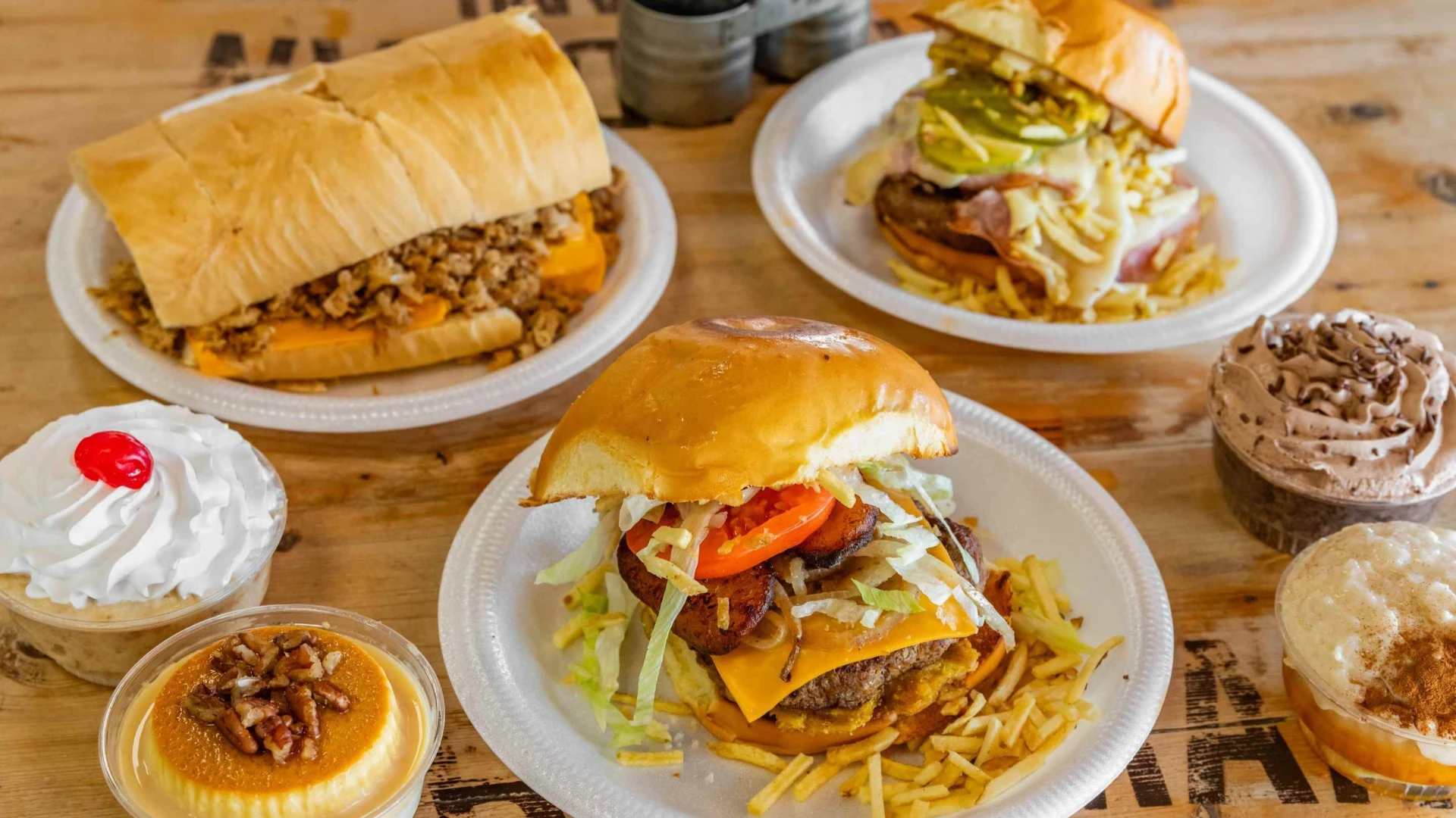 Assorted Breadman Miami dishes including loaded sandwiches, burgers with cheese, plantains, and shredded lettuce, served with sides of flan, whipped cream dessert, chocolate mousse, and rice pudding on a wooden table