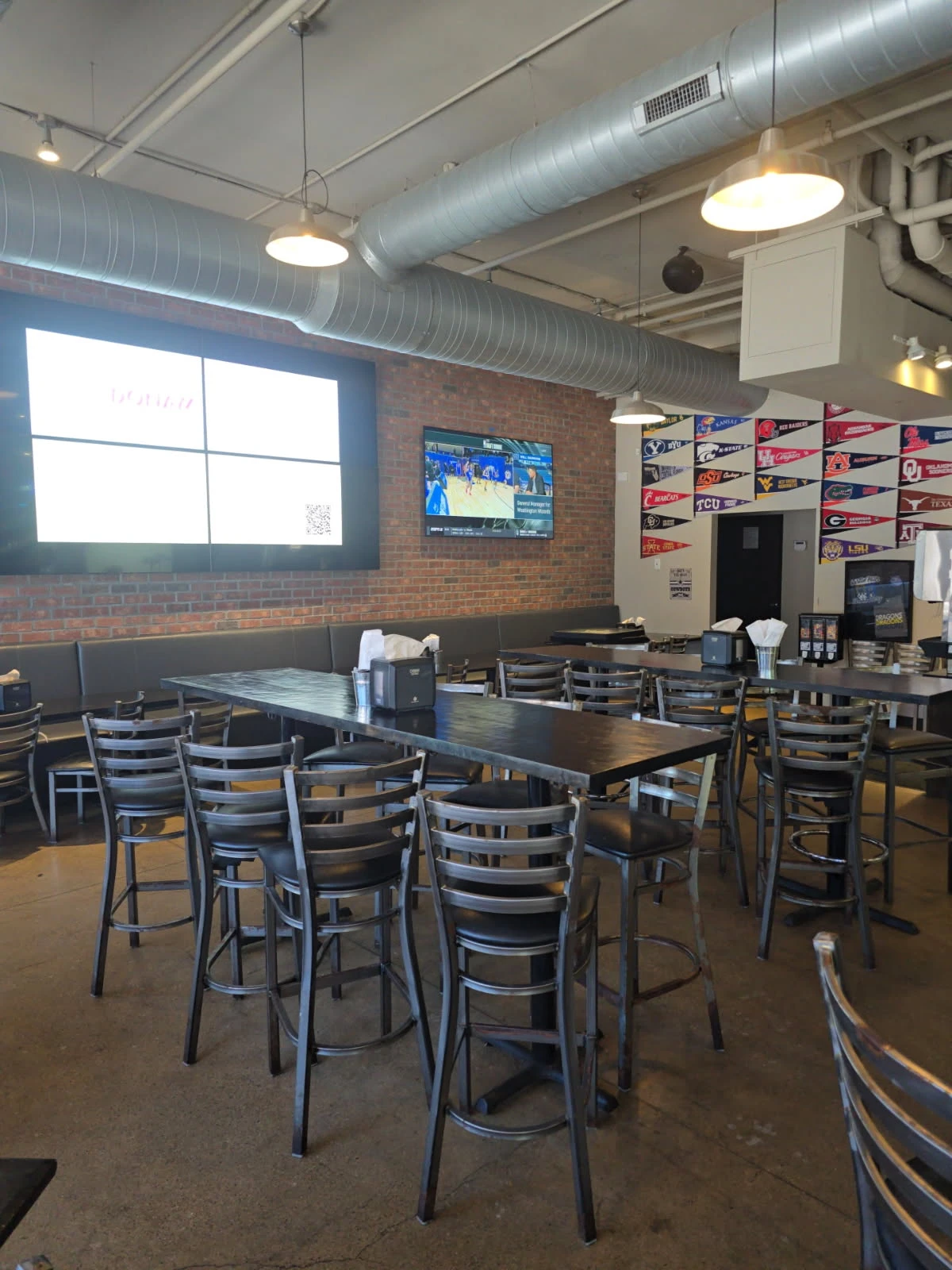 Indoor restaurant seating area with large TV screens, brick walls, and college pennant decor