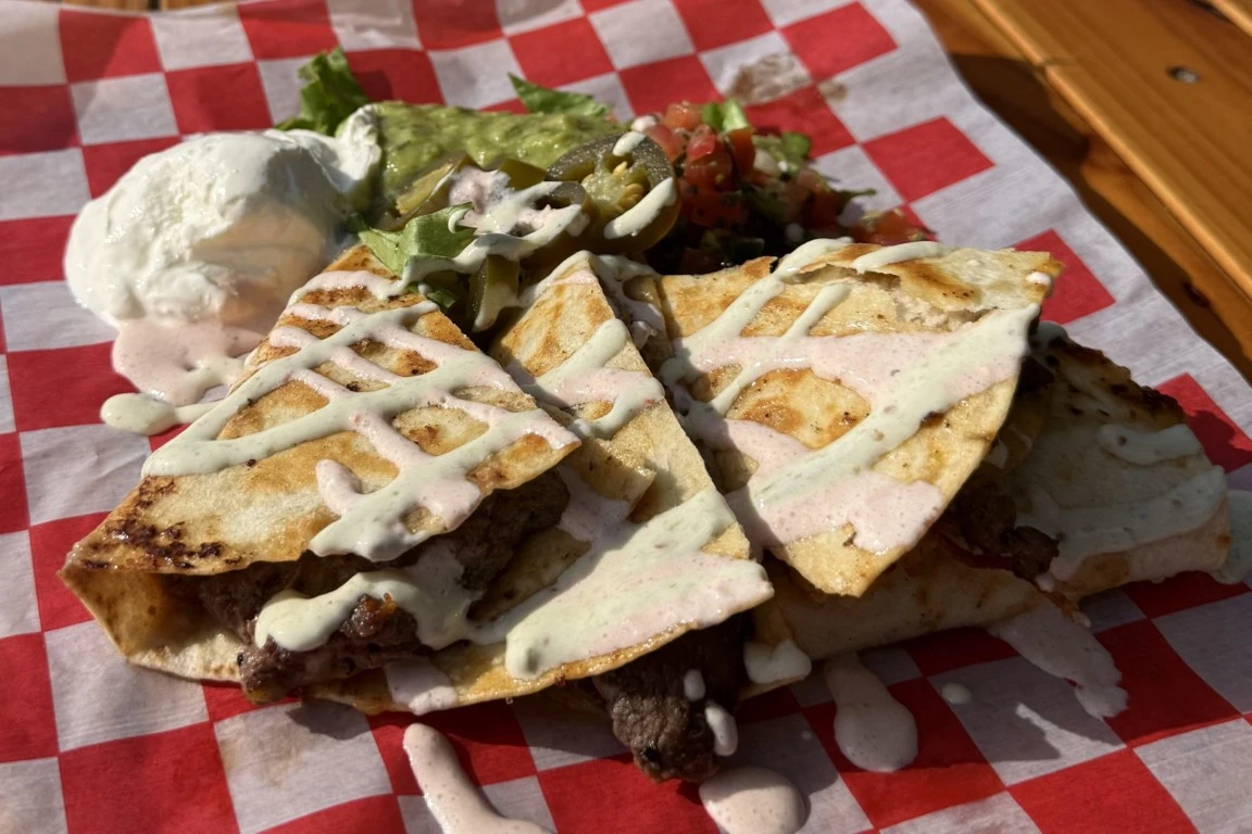 Grilled steak quesadilla wedges topped with creamy sauce, served with sour cream, guacamole, jalapeños, pico de gallo, and lettuce on a red checkered tray liner