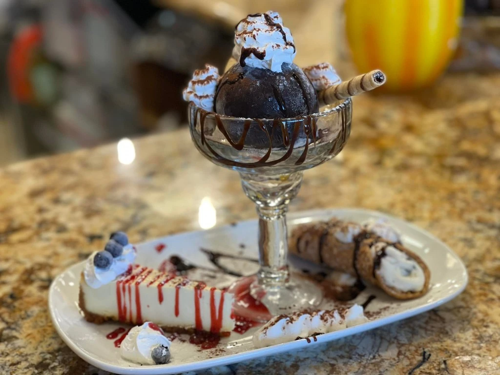 Dessert platter featuring a chocolate gelato sundae in a glass dish, a slice of cheesecake with berry sauce and whipped cream, and a classic cannoli on a rectangular plate.