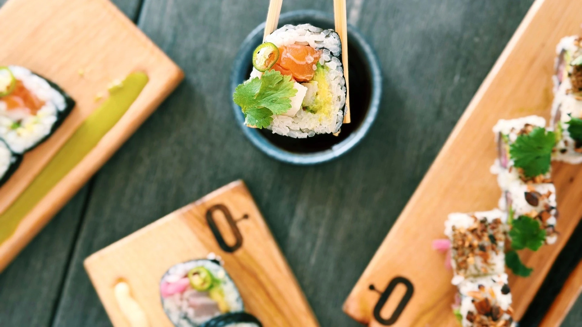 Close-up of a fresh sushi roll held with chopsticks above a dipping sauce dish, with assorted sushi rolls on wooden boards.