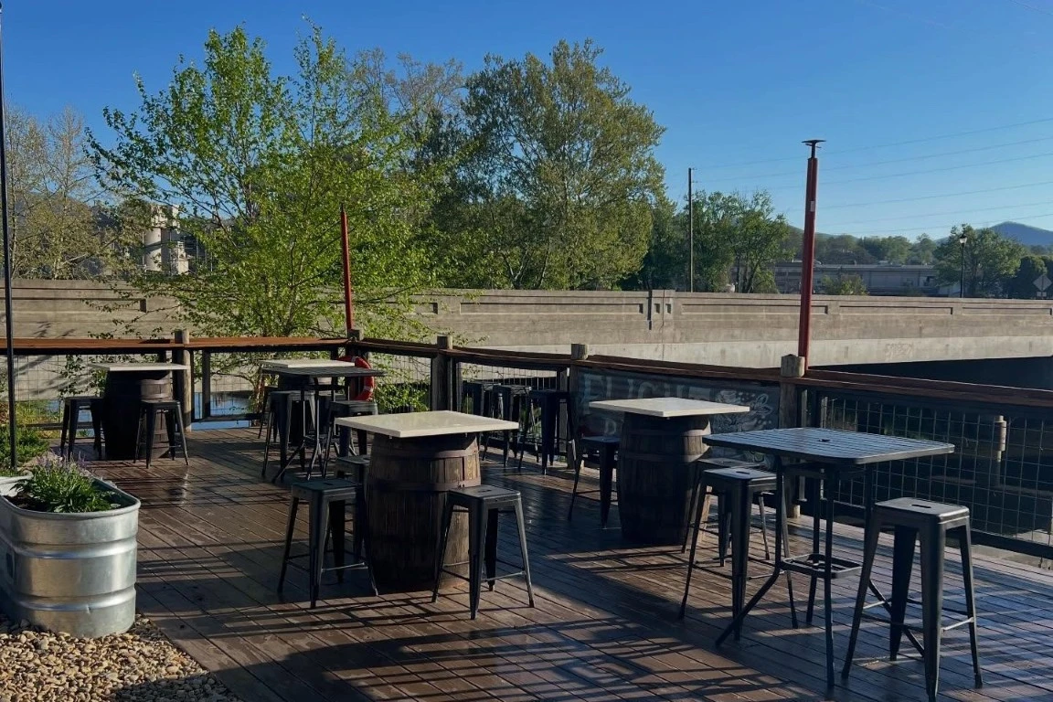 Outdoor rooftop patio with high-top tables and stools on a wooden deck overlooking trees and a nearby river on a clear sunny day.