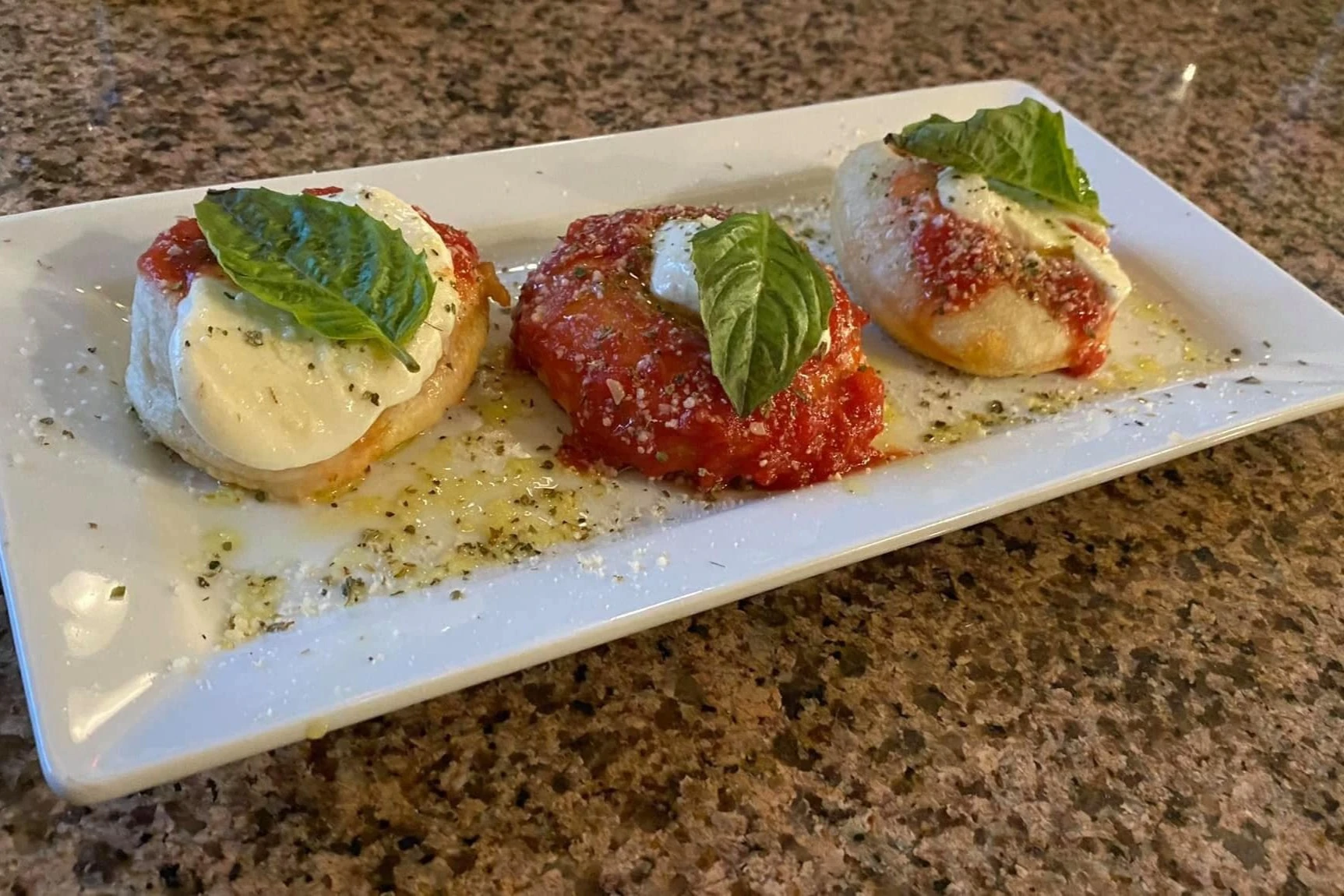 Three fried dough rounds topped with fresh mozzarella, tomato sauce, grated cheese, and basil leaves served on a rectangular white plate.