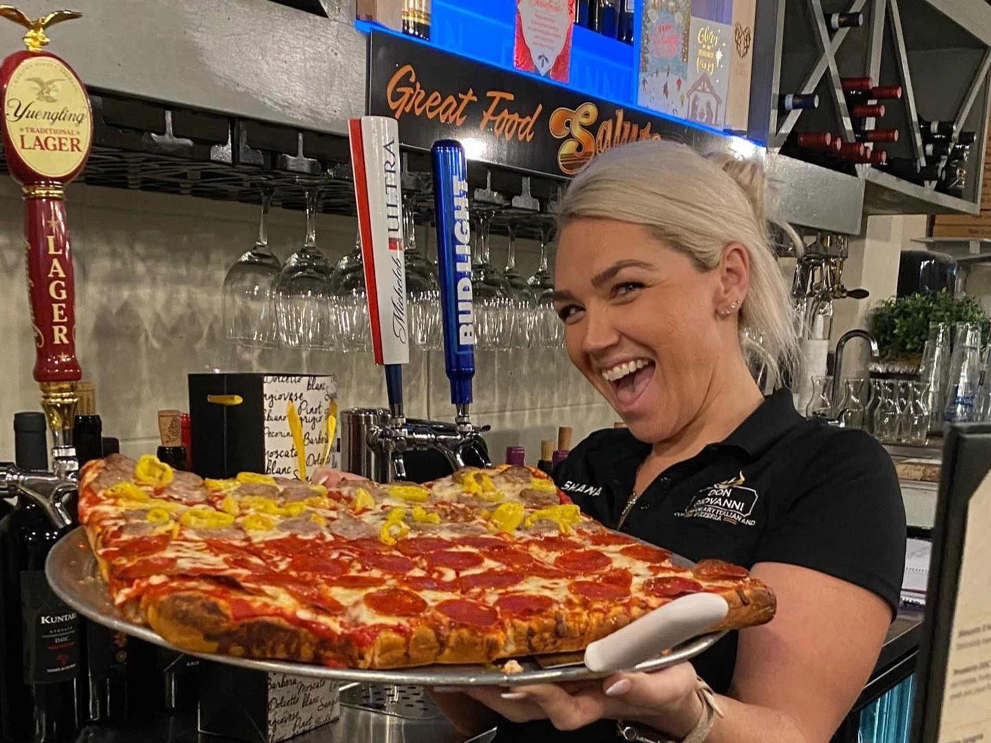 Smiling restaurant staff member holding a large rectangular pizza topped with pepperoni and banana peppers behind the bar area.