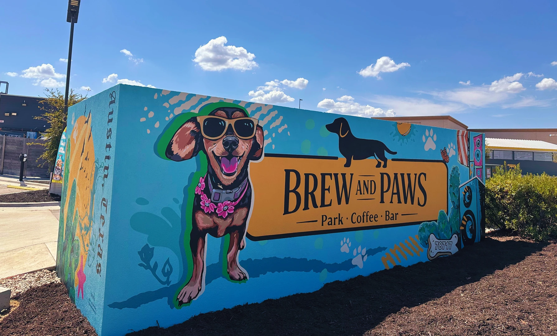 A colorful outdoor mural features a dog wearing sunglasses beside the text “BREW AND PAWS Park · Coffee · Bar”