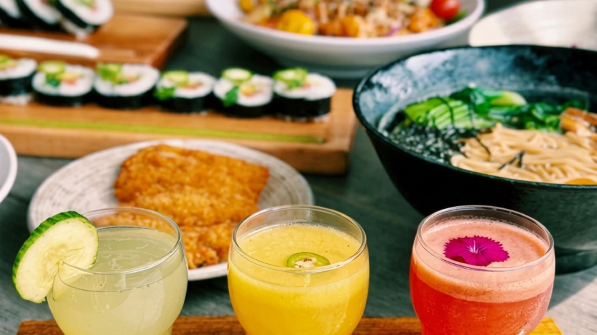 Colorful sushi rolls, ramen bowl, crispy entrée, and handcrafted cocktails arranged on a restaurant table.