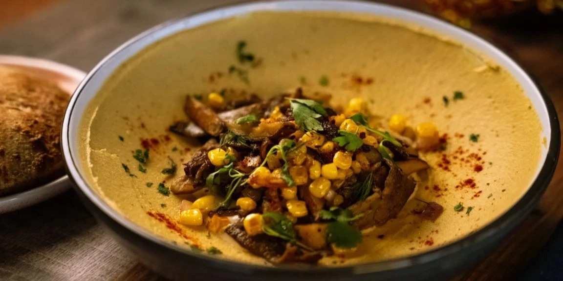 Creamy hummus bowl topped with sautéed mushrooms, corn, fresh herbs, and paprika, served with warm bread on the side.