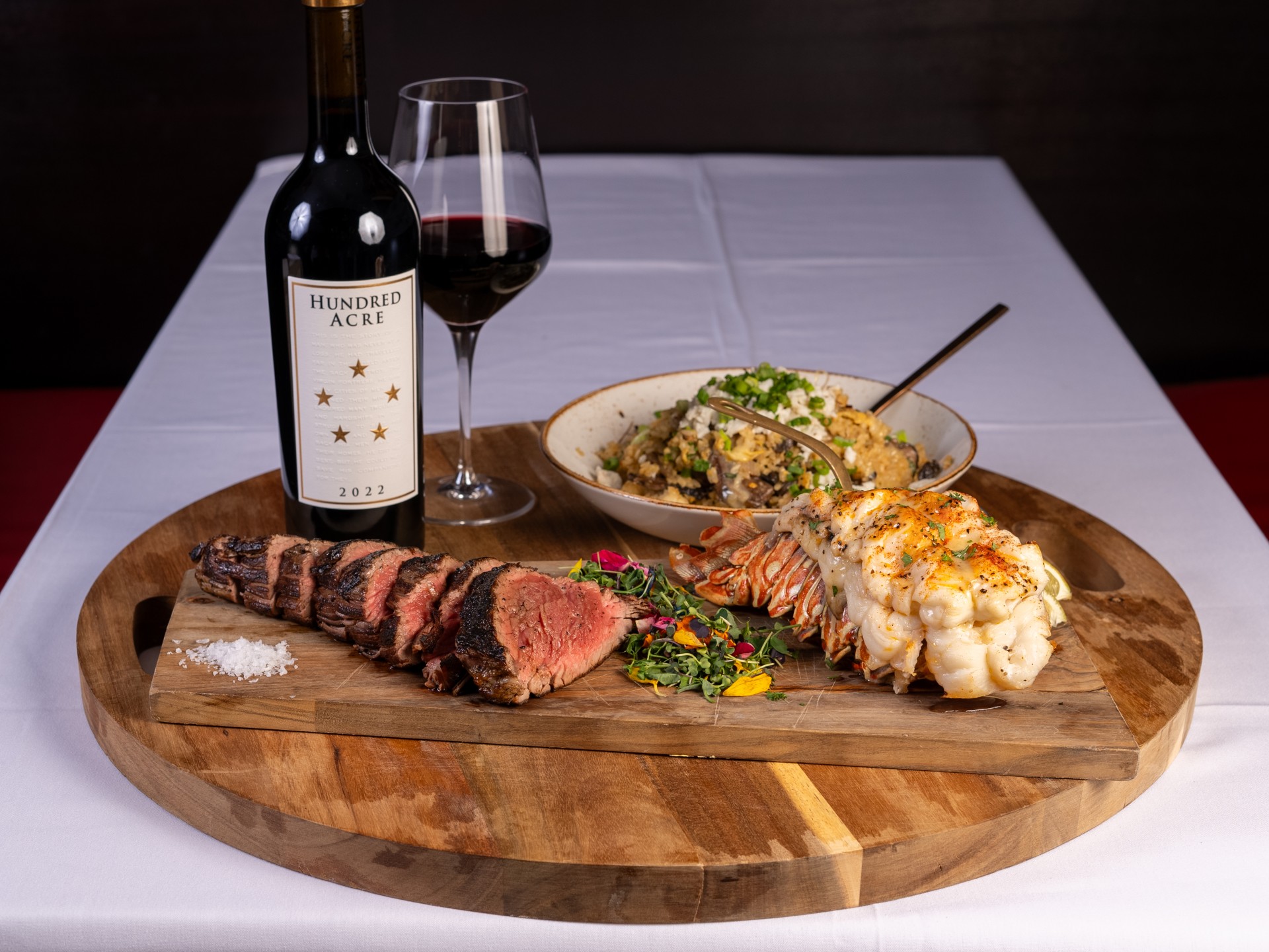 Steak and lobster on a serving tray with wine and sides in the background