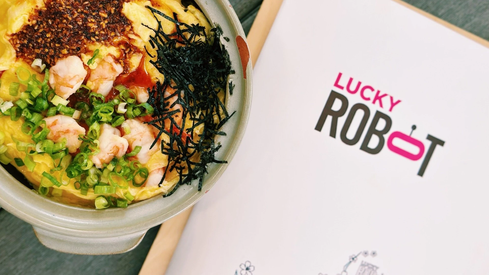 Japanese-style egg and shrimp rice bowl topped with green onions and seaweed beside a Lucky Robot menu.
