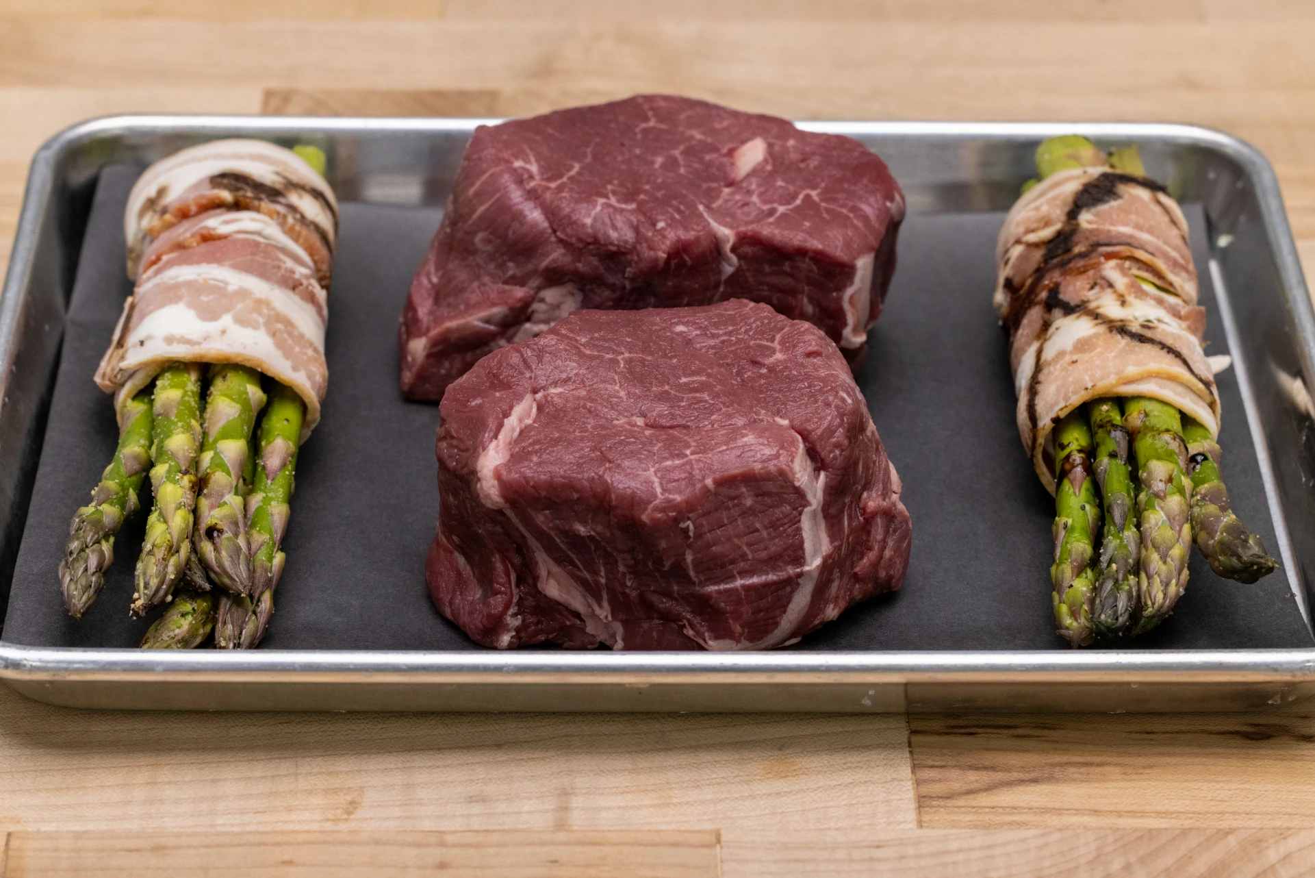 Raw beef filet cuts and bacon-wrapped asparagus arranged on a tray