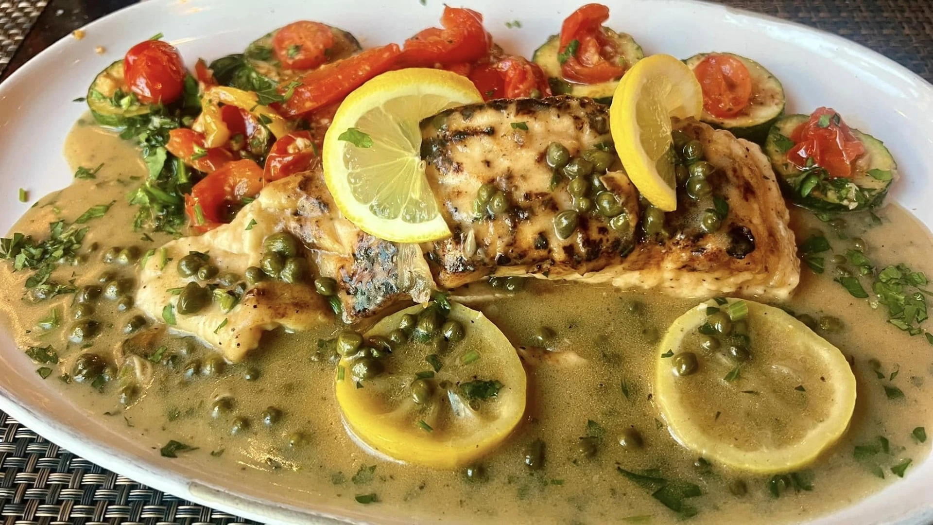Grilled fish piccata with lemon slices, capers, and a vegetable medley in a buttery sauce