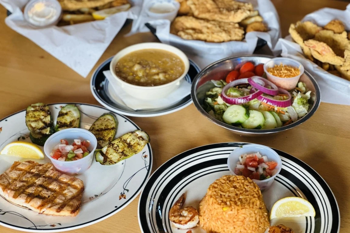 Table of plates featuring grilled salmon, grilled zucchini, seasoned rice, pico de gallo, a fresh garden salad, bowls of soup, and baskets of fried seafood.