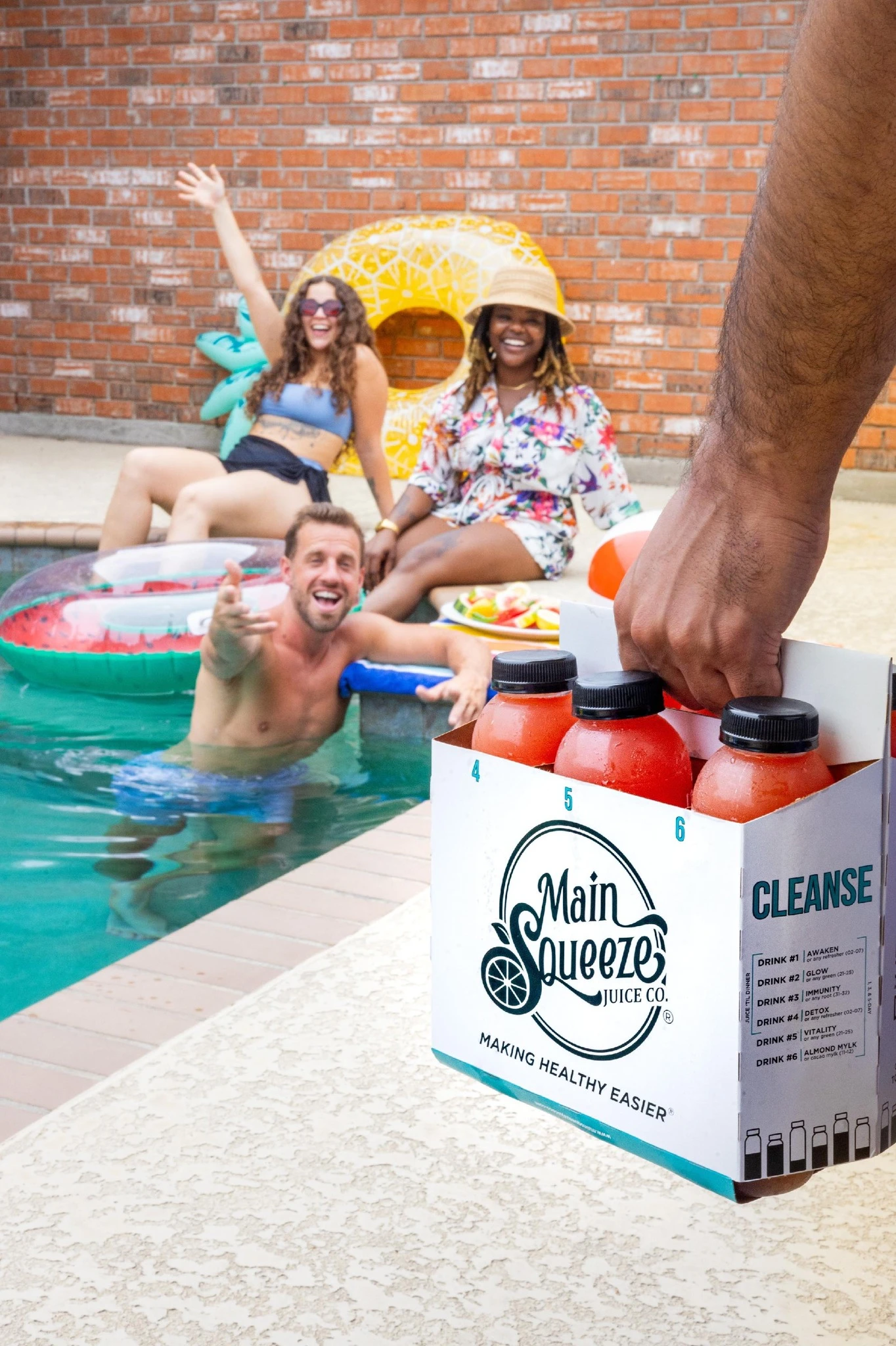 Group of friends by the pool with a Main Squeeze Juice Co. cleanse pack of juices in the foreground