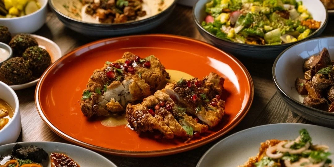 Crispy fried chicken topped with pomegranate seeds and herbs on an orange plate, surrounded by assorted Mediterranean sides including salads, falafel, and roasted vegetables.