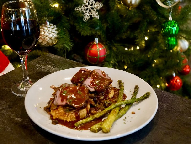 A plated holiday entrée of sliced roast with rich sauce and roasted asparagus, Christmas tree in the background