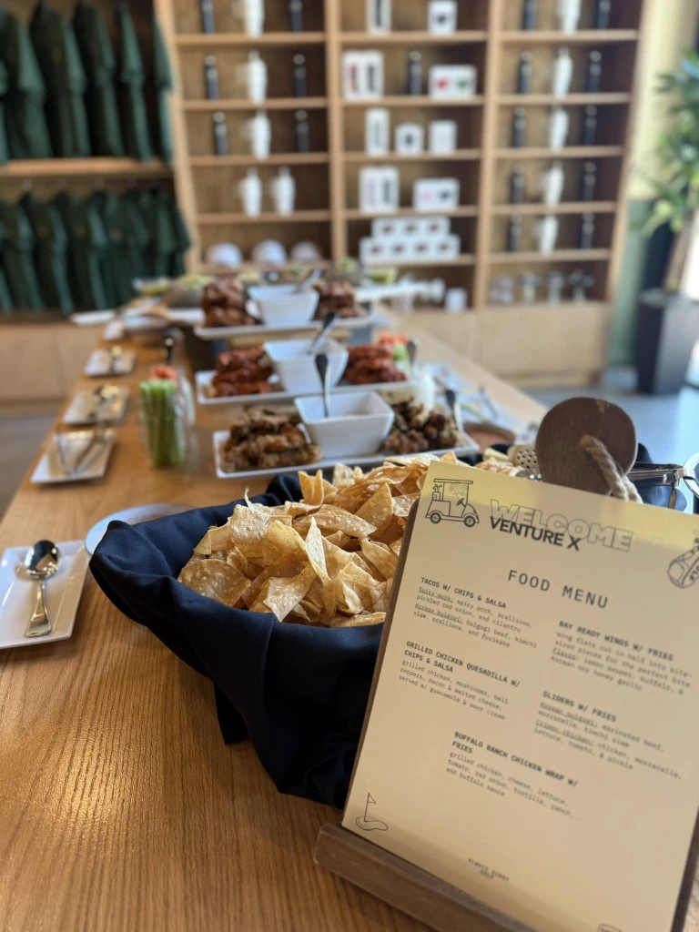 Catering spread with tortilla chips, tacos, wings, and event menu display on a wooden table inside a modern venue