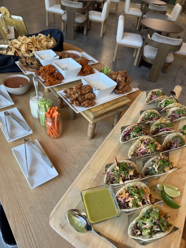 Buffet-style catering table featuring tacos, chicken wings, chips, vegetables, dips, and sauces arranged on serving platters
