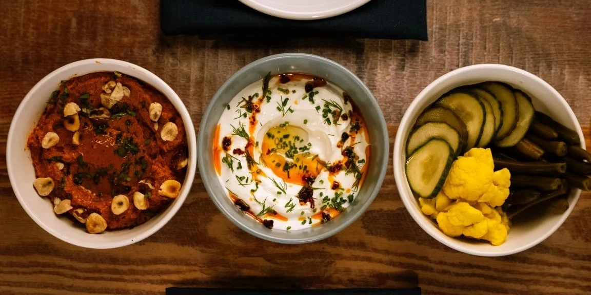 Three Mediterranean bowls on a wooden table with spicy red dip topped with hazelnuts, creamy labneh drizzled with oil and herbs, and assorted pickled vegetables including cucumbers, cauliflower, and green beans.