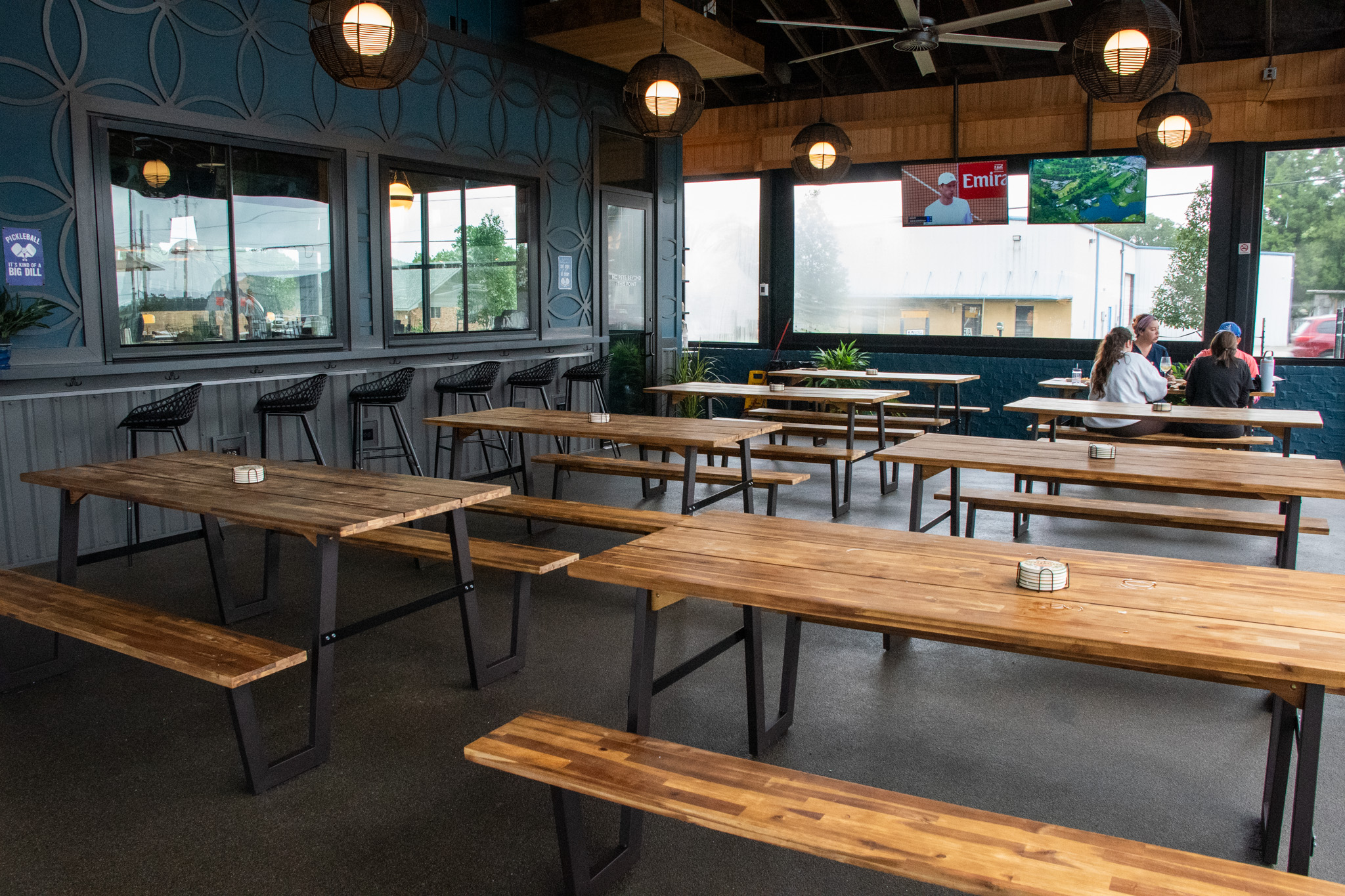 Covered patio with long wooden communal tables, pendant lights, and TVs
