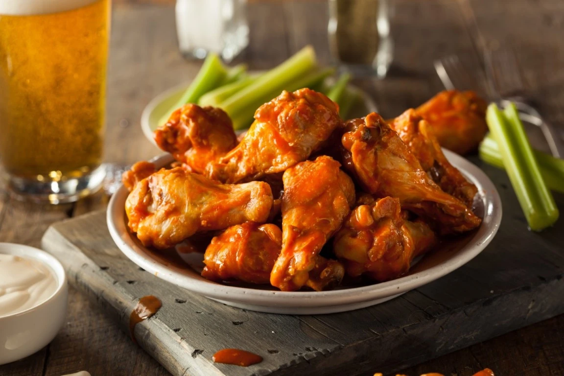 Plate of buffalo chicken wings with celery sticks, ranch dipping sauce, and beer in the background