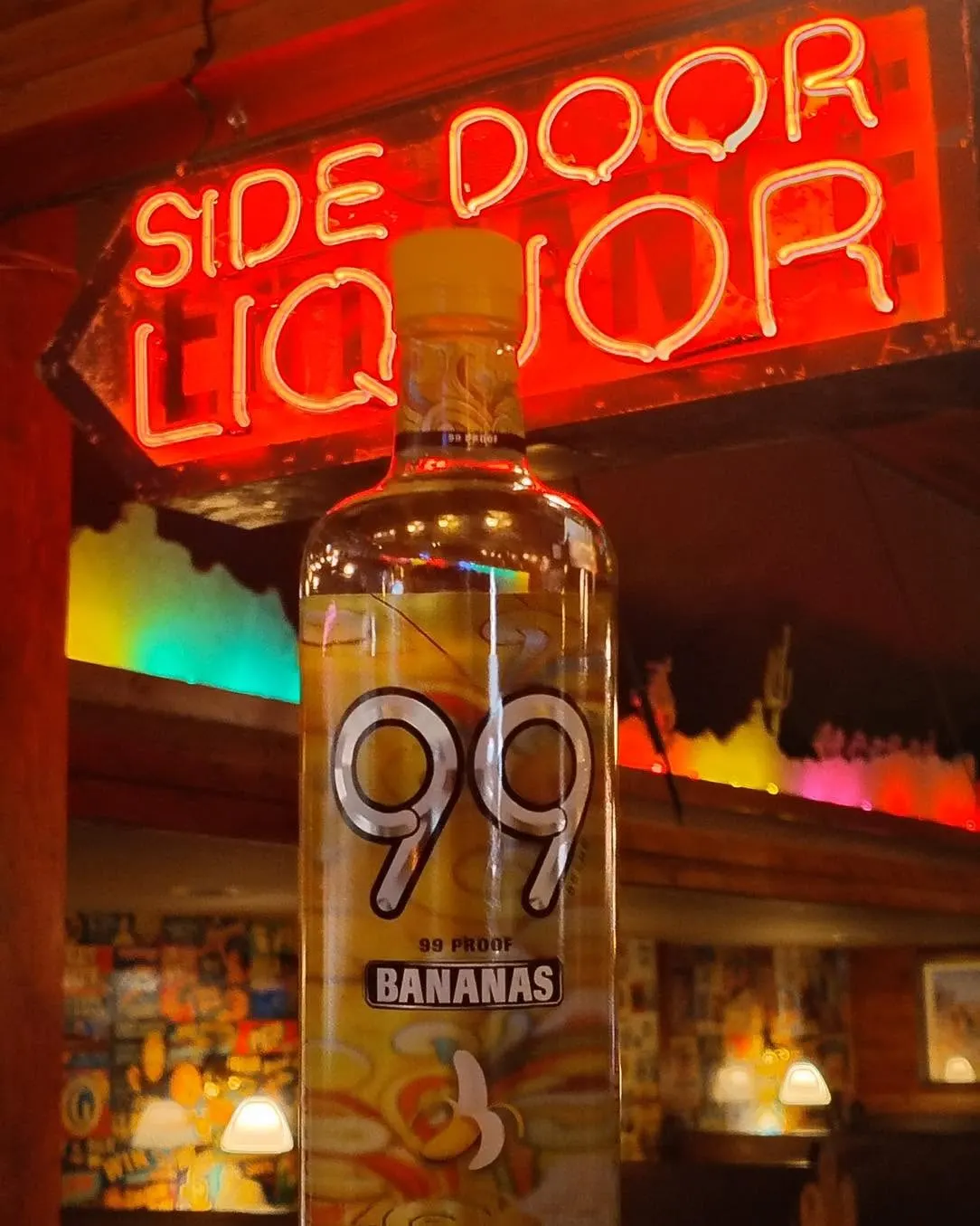 A bottle of 99 Bananas held up beneath the red Side Door Liquor neon sign inside the store