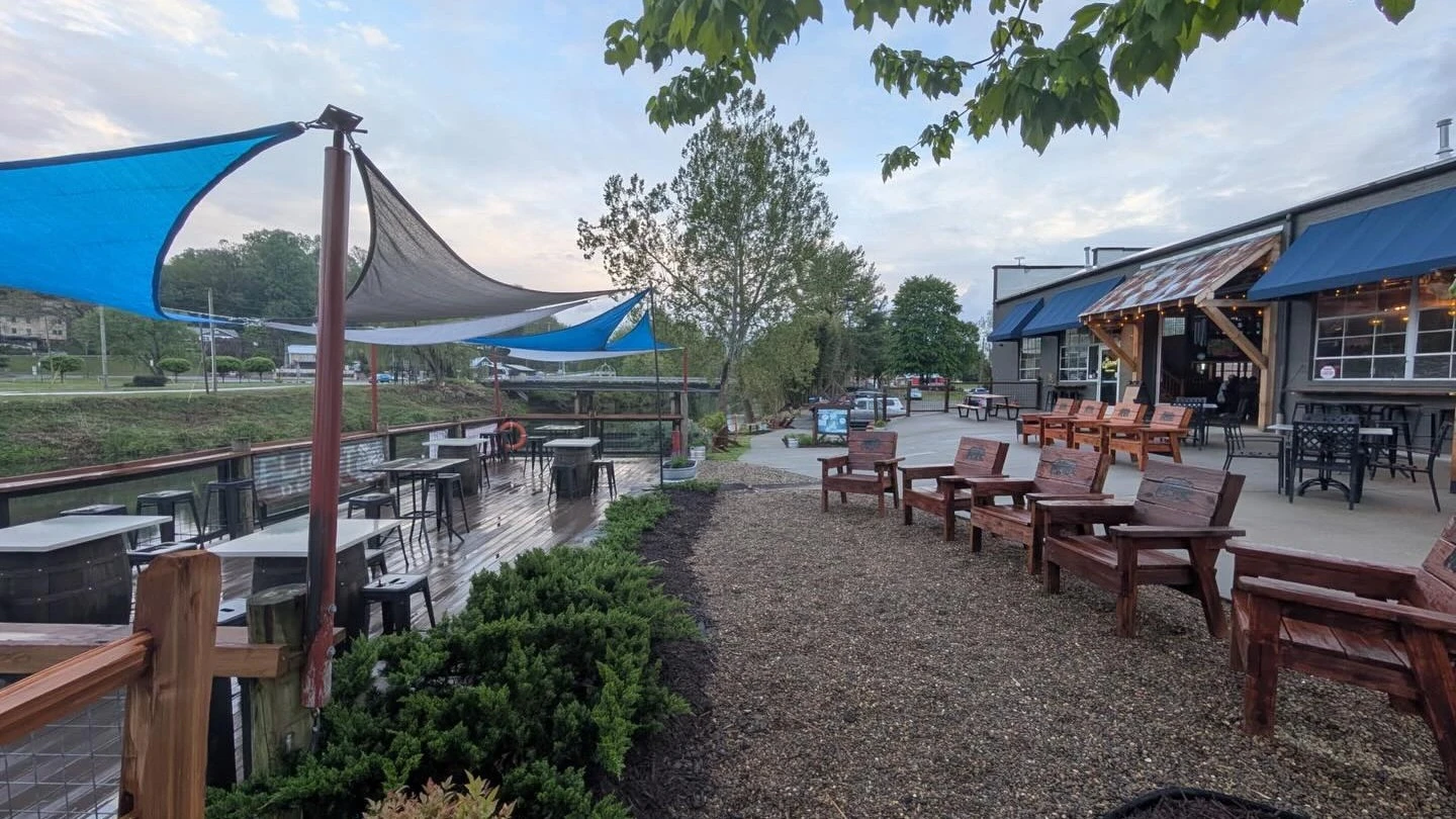 Outdoor riverside patio seating area with shade sails, wooden chairs, and tavern building exterior