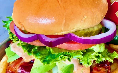 Burger on a toasted bun with red onion, tomato, pickles, lettuce, avocado, bacon and a creamy sauce