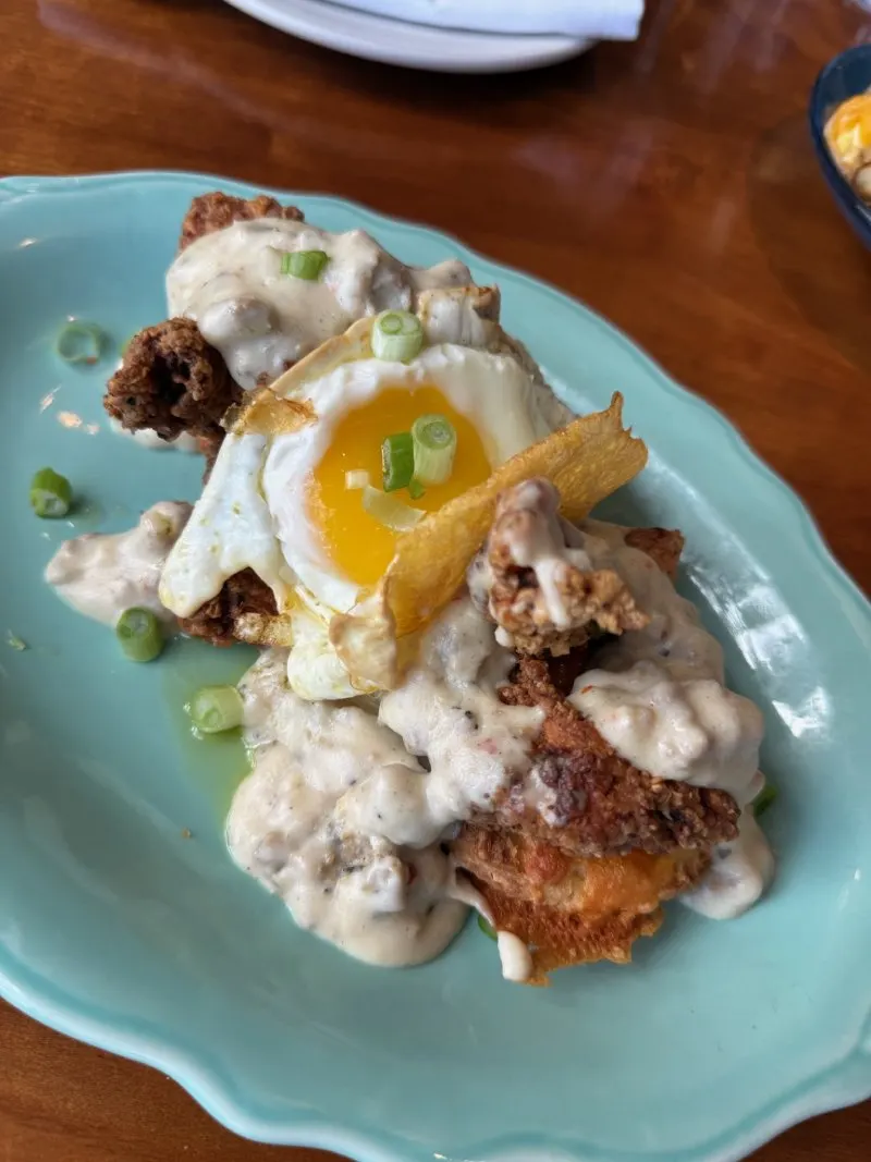 Chicken biscuits smothered in gravy and topped with a soft egg, served on a blue plate at Junior’s on Harrison