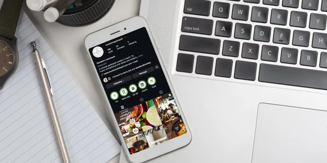Smartphone displaying an Instagram profile on a desk next to a laptop keyboard, notebook, pen, watch, and camera lens