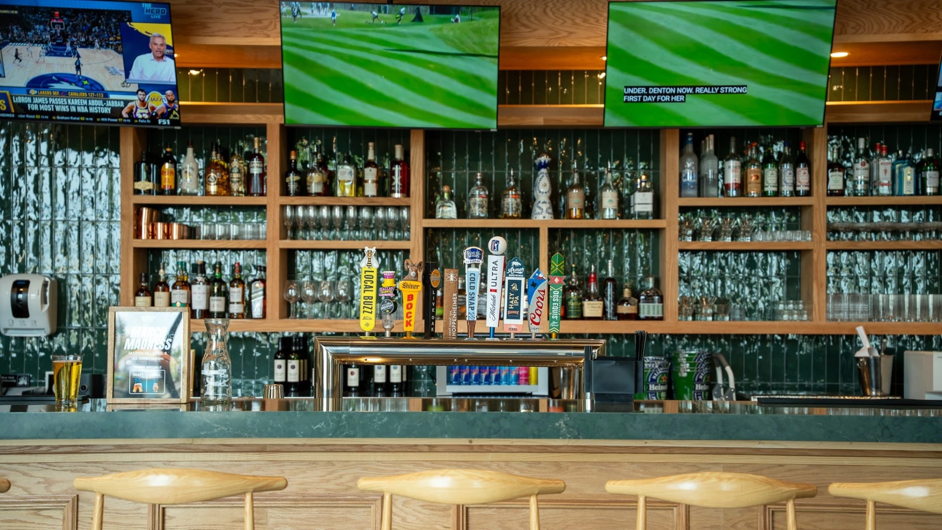 Sports bar with liquor display, beer taps, large TVs showing golf coverage, and wood-accented shelving
                                                                                                                                               