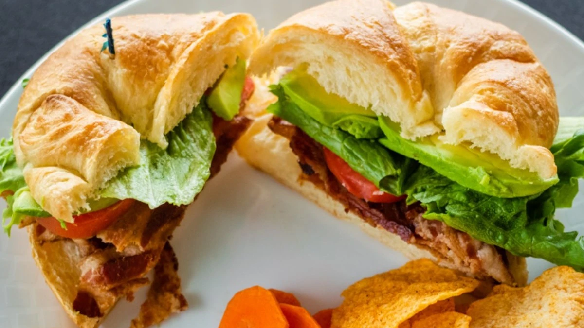 Croissant sandwich filled with lettuce, tomato, bacon, and avocado served with chips and sliced carrots