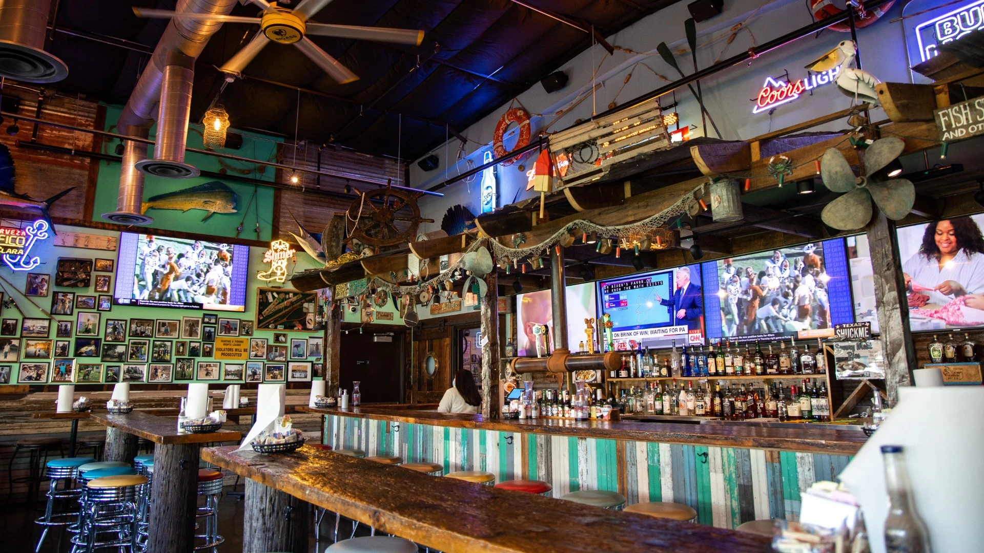 olorful bar interior with rustic nautical decor, neon signs, mounted TVs, and a long wooden bar with stools.