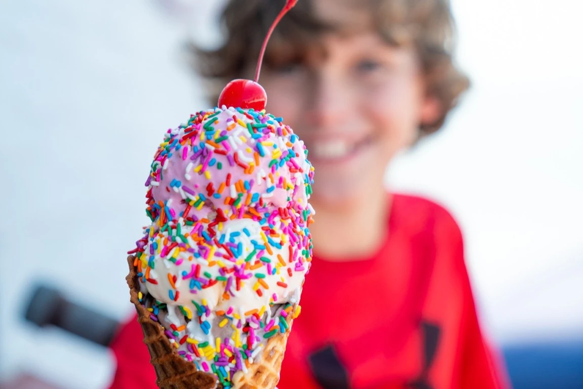 A kid holding a double scoop of ice cream with sprinkles and cherry on top in a waffle cone