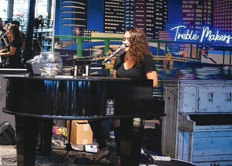 a person playing a grand piano and singing with a blue neon sign saying Table Makers in the background