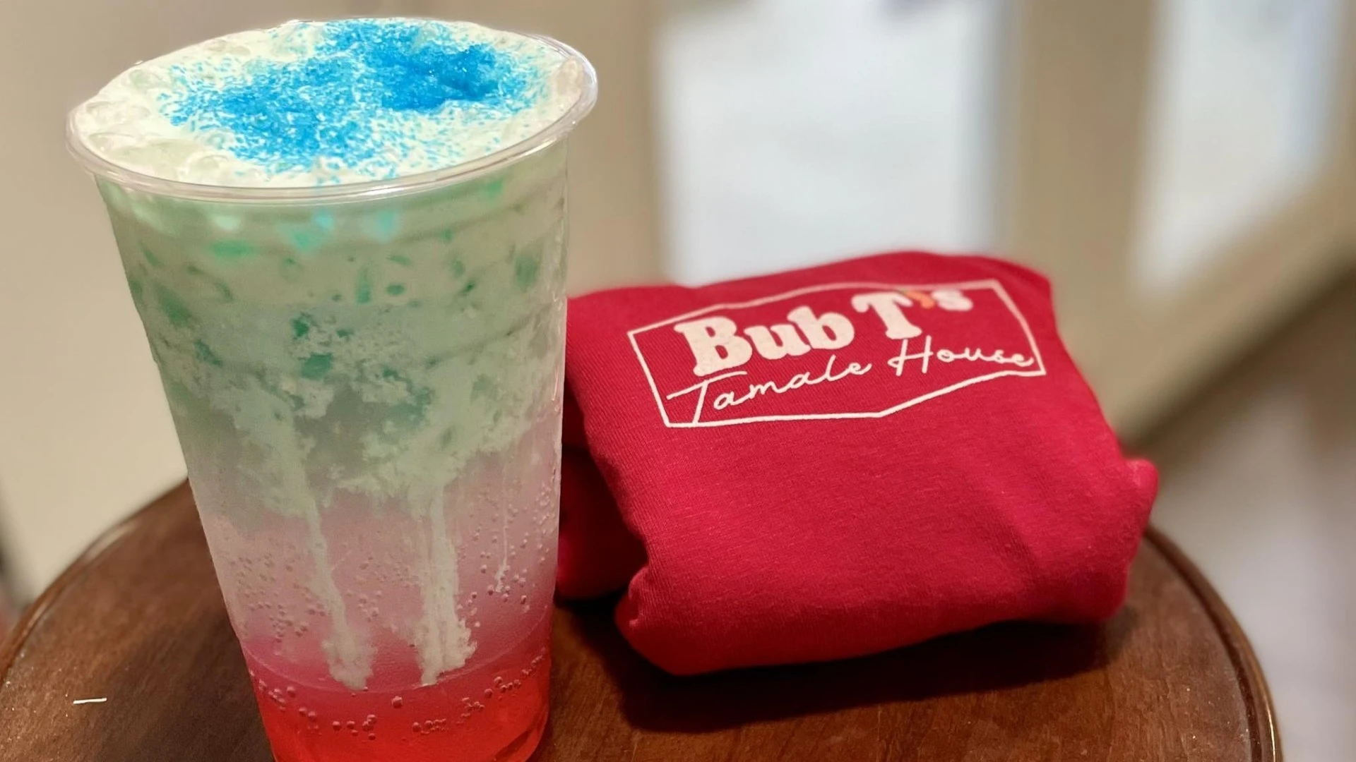 Red and blue layered frozen drink in a clear cup next to a Bub T's Tamale House branded cloth on a wooden tray